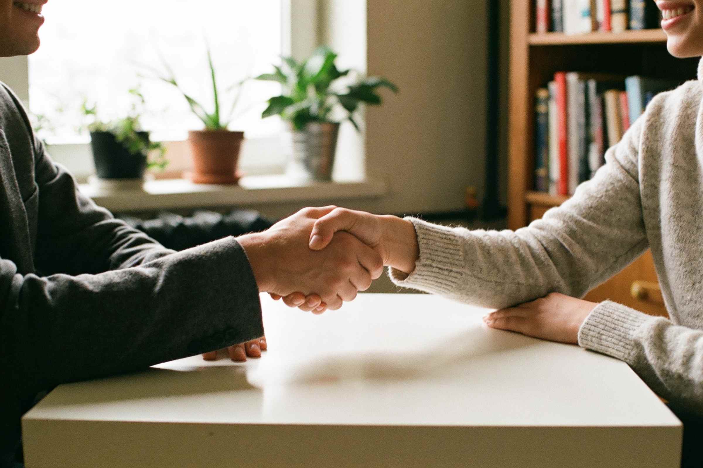 Two people shaking hands across a desk in a small bright office